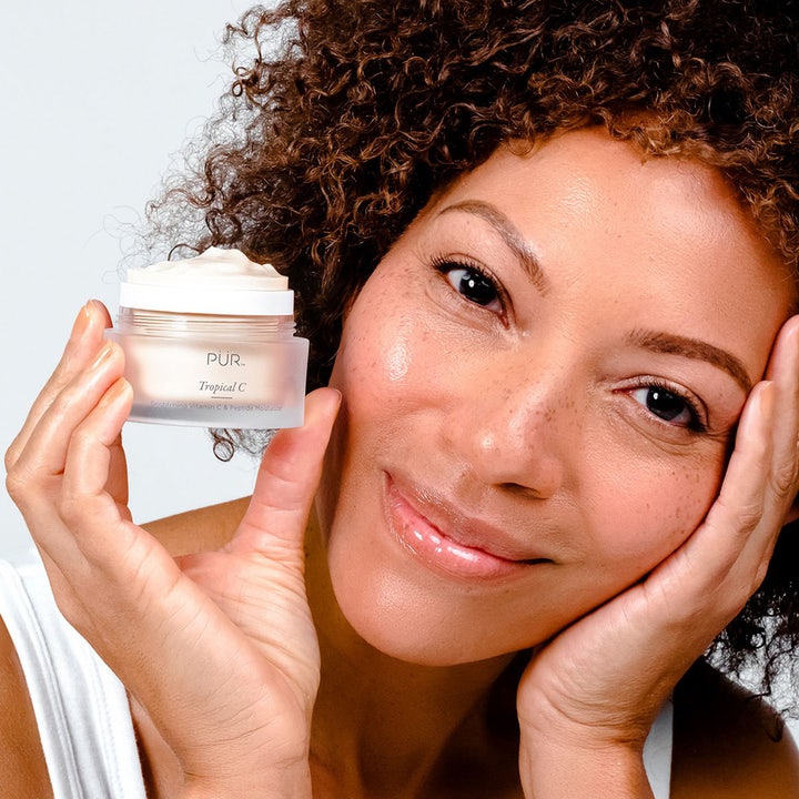 PÜR Tropical C Vitamin C Moisturizer 50 ml i glasskrukke holdt av smilende kvinne med glødende hud