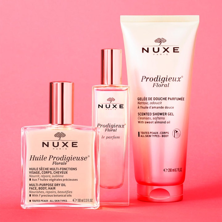 Nuxe Huile Prodigieuse Floral tørrolje i glassflaske sammen med parfyme og dusjgelé på en rosa bakgrunn.