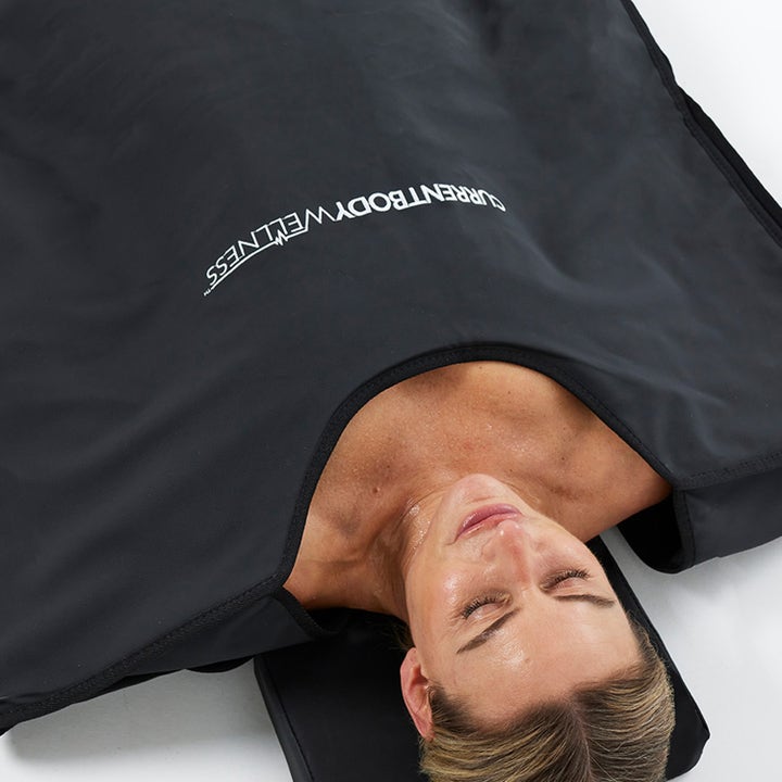 Currentbody Skin Sauna Blanket infrarødt varmeteppe for restitusjon med kvinne som ligger i det svarte teppet.