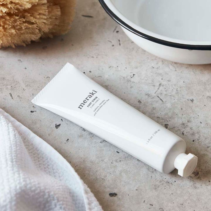 Meraki Footscrub Calm Cedar 100 ml eksfolierende fotpleie i hvit tube som mykgjør huden ved et vaskefat.