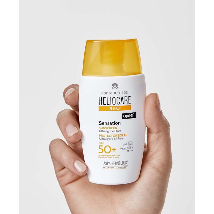 Heliocare Sensation SPF 50+ 50 ml lett solkrem for ansiktet i hvit flaske med gult lokk som holdes i en hånd