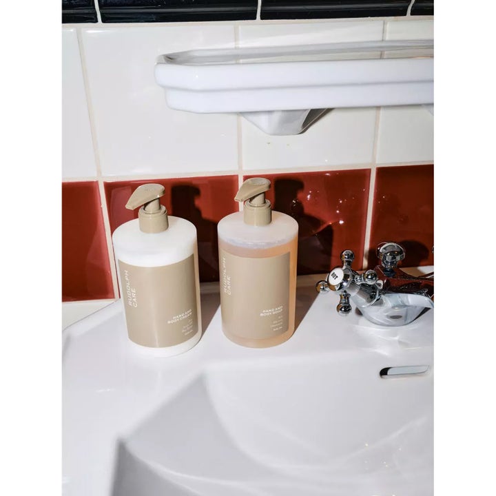 Rudolph Care Hand And Body Soap 500 ml pleiende dusjsåpe i beige pumpeflaske på en hvit vask ved en kromkran.