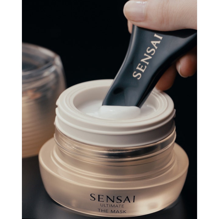 Sensai Ultimate The Mask 75 ml ansiktsmaske i gullfarget krukke med en svart spatel og hvit kremet tekstur