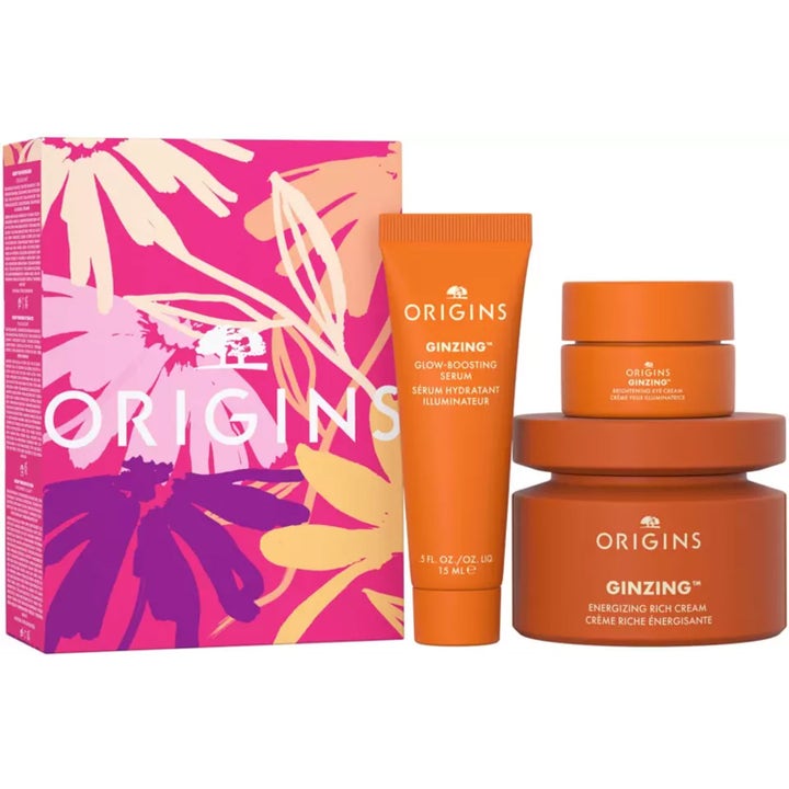 Origins GinZing 3 stk energigivende hudpleiesett som gir glød med oransje krukker og tube ved en rosa blomstret eske.