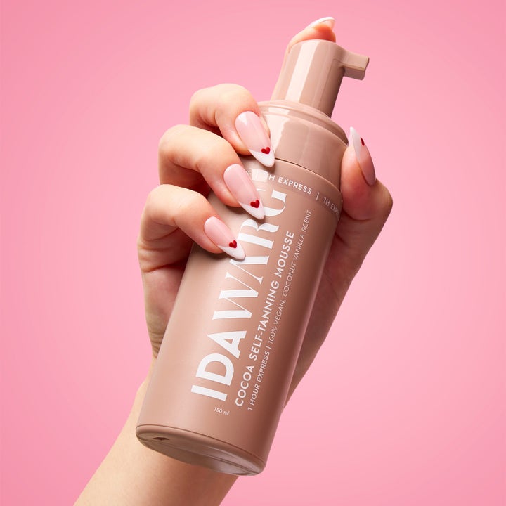IDA WARG Cocoa Self-Tanning Mousse 150 ml gir rask glød i en brun pumpeflaske holdt av en hånd mot rosa bakgrunn.