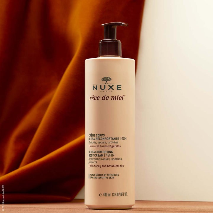 Nuxe Rêve de Miel Comforting Body Cream 400 ml fuktighetsgivende body lotion i beige pumpeflaske mot varm bakgrunn