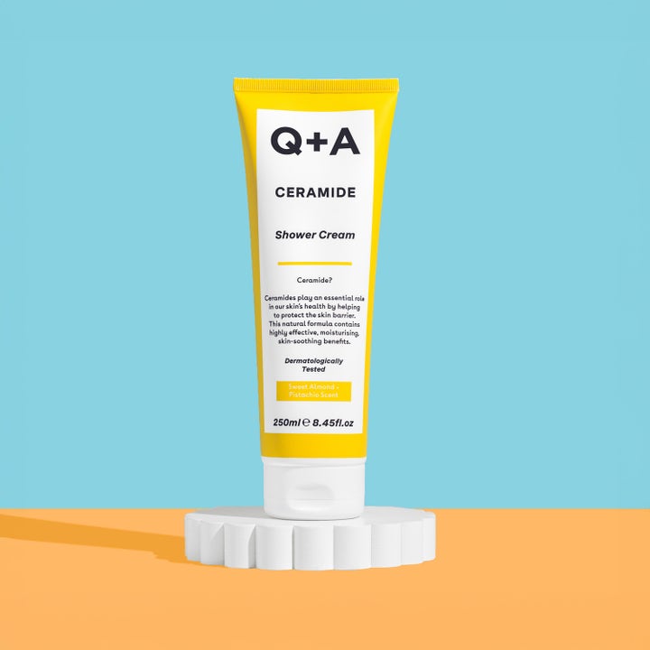 Q+A Ceramide Shower Cream 250 ml fuktighetsgivende dusjsåpe i gul tube på hvit sokkel mot fargerik bakgrunn.