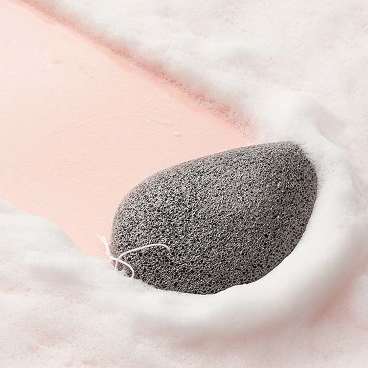 Purito Bamboo Charcoal Konjac Sponge 7 g eksfolierende rengjøringssvamp i grått omgitt av hvitt skum på rosa bakgrunn
