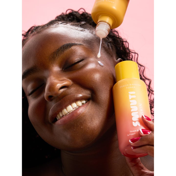 Smuuti Skin Peach Barrier Toner 130 ml ansiktsvann gir glød mens dråper påføres huden til en smilende kvinne.