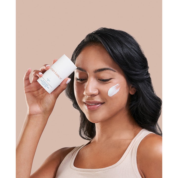Exuviance Overnight Transformation Complex 50 g nattkrem i hvit flaske vist på en kvinne med krem på kinnet.