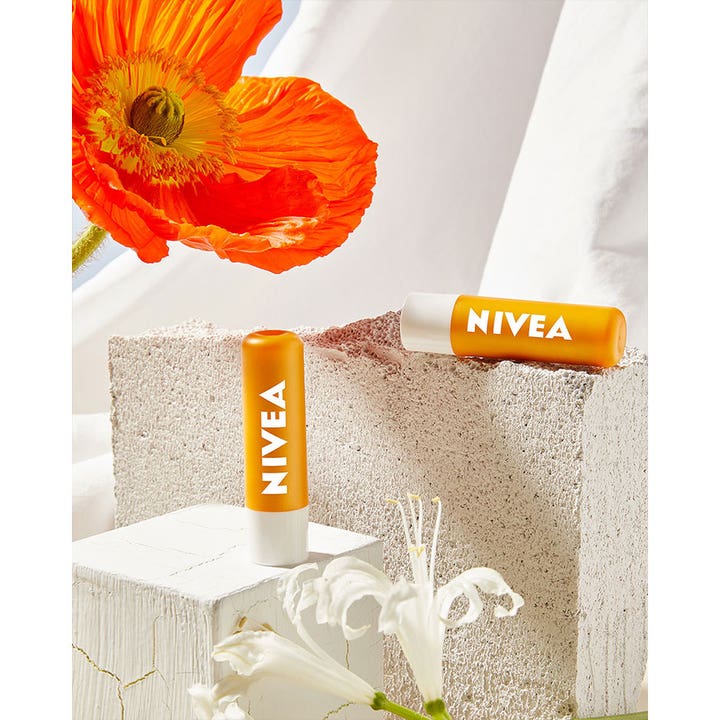 NIVEA Sun Protect Sunstick 4 g gir solbeskyttelse i oransje hylser plassert på hvite steiner ved en oransje blomst