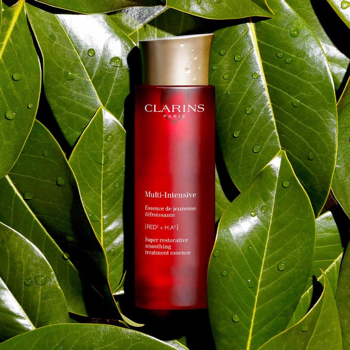 Clarins Super Restorative Treatment Essence 200 ml utglattende essence i rød flaske på grønne blader med vanndråper