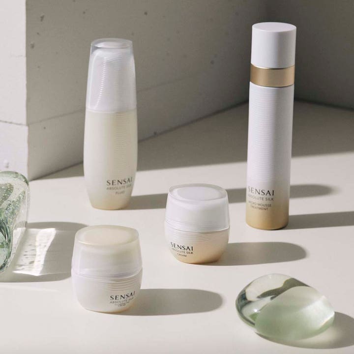 Sensai Absolute Silk Illuminative Cream 40 ml ansiktskrem gir glød i hvit krukke ved siden av andre produkter.
