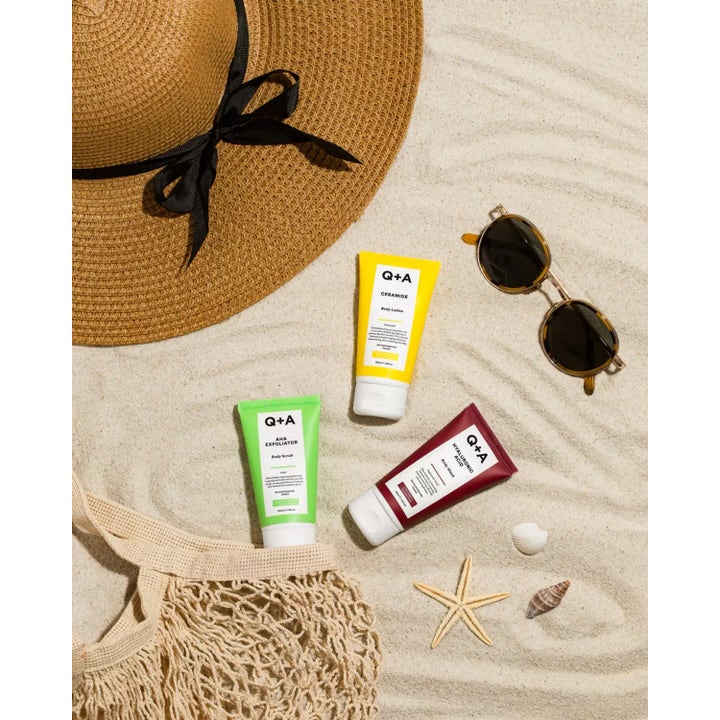 Q+A AHA Exfoliator Body Scrub 50 ml eksfolierende kroppsskrubb i grønn tube på sand med solhatt og solbriller