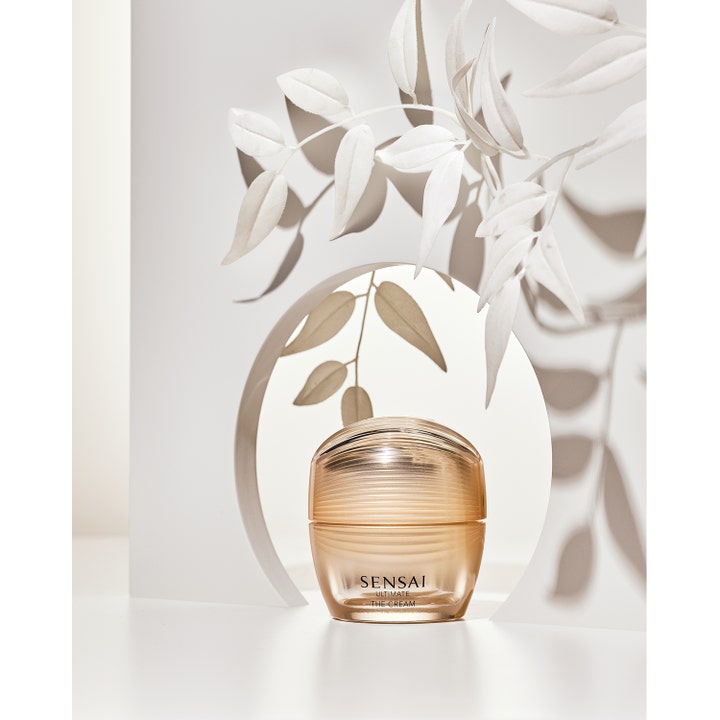 Sensai Ultimate The Cream 40 ml ansiktskrem i gyllen krukke foran en hvit bue med blader og skygger.