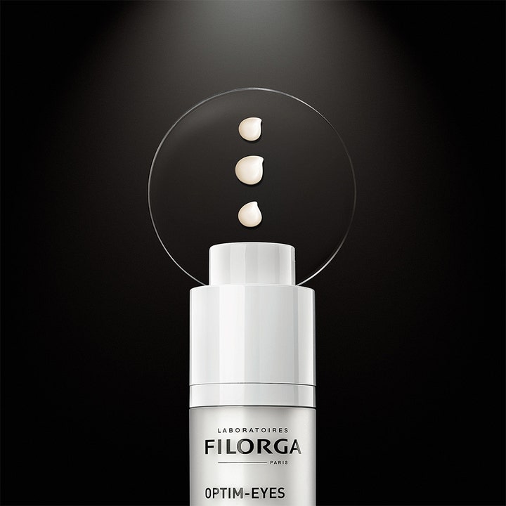 Filorga Optim-Eyes 15 ml øyekrem mot mørke ringer viser tre lyse kremdråper over en hvit pumpeflaske