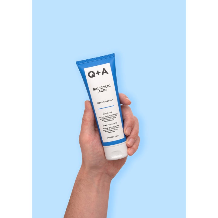 Q+A Salicylic Acid Cleanser 125 ml eksfolierende ansiktsrengjøring i blå tube som holdes i en hånd mot blå bakgrunn.