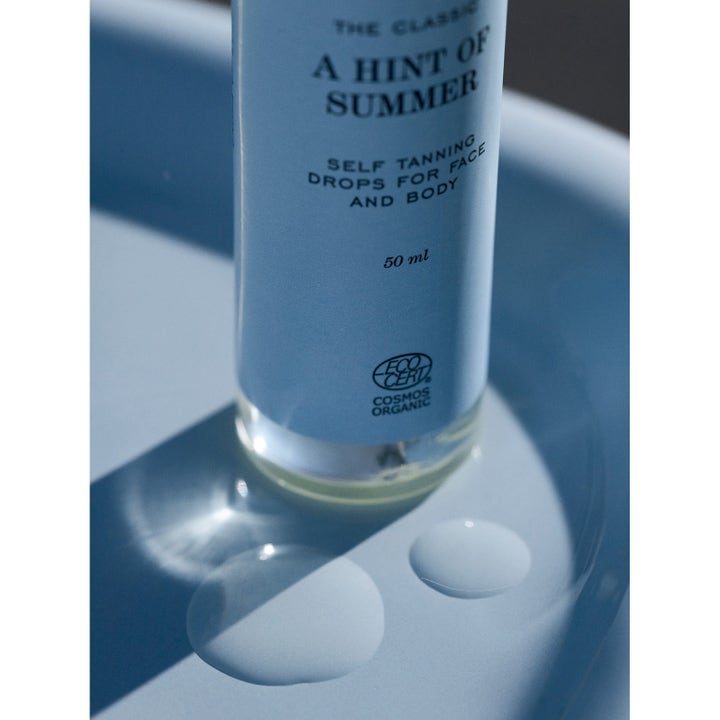 Rudolph Care A Hint of Summer 50 ml selvbruningsdråper som gir glød vist med klare dråper på en blå flate.