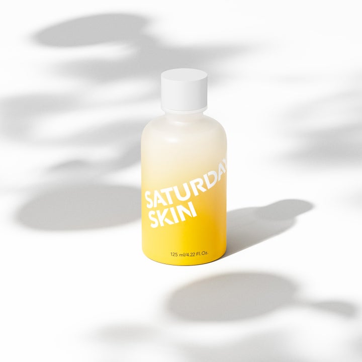 Saturday Skin Yuzu Vitamin C Brightening Toner 125 ml gir glød i en gul og hvit frostet flaske omgitt av skygger.