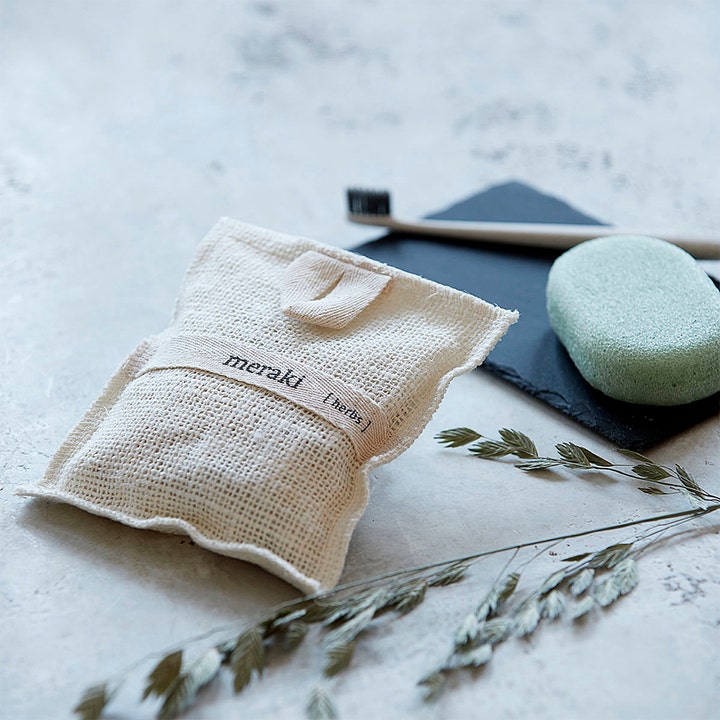 Meraki Bath Mitt Herbs 140 g beroligende badeprodukt i beige tekstilpose ved en grønn såpe og tørkede urter.