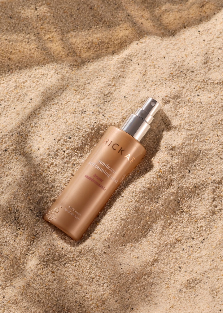 Hickap Sundazed Self Tanning Mist 100 ml gir glød i en bronsefarget sprayflaske som ligger på sand.