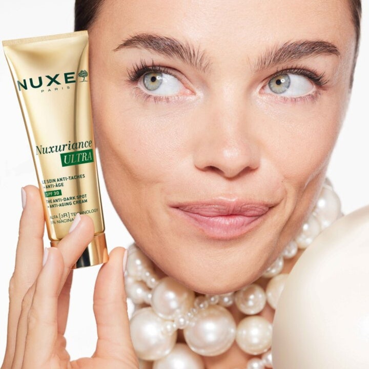 Nuxe Nuxuriance Ultra Anti Dark Spot Cream 50 ml dagkrem med SPF 30 i gulltube holdt av kvinne med perlekjede