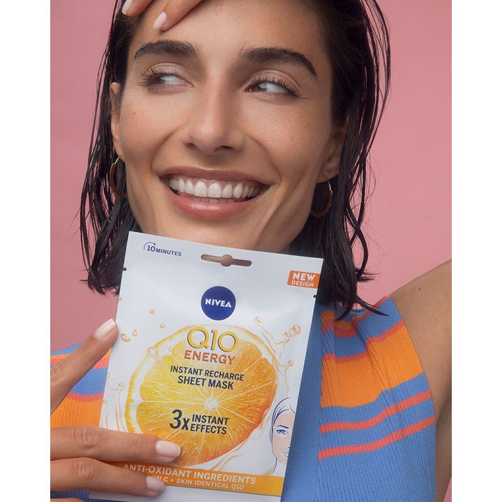NIVEA Q10 Energy Sheet Mask 1 stk glødgivende ansiktsmaske med Vitamin C holdt av smilende kvinne mot rosa bakgrunn