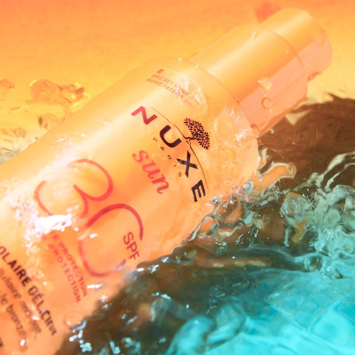 Nuxe Sun Spray SPF30 184 g solbeskyttelse i en oransje sprayflaske som ligger i glitrende blått vann