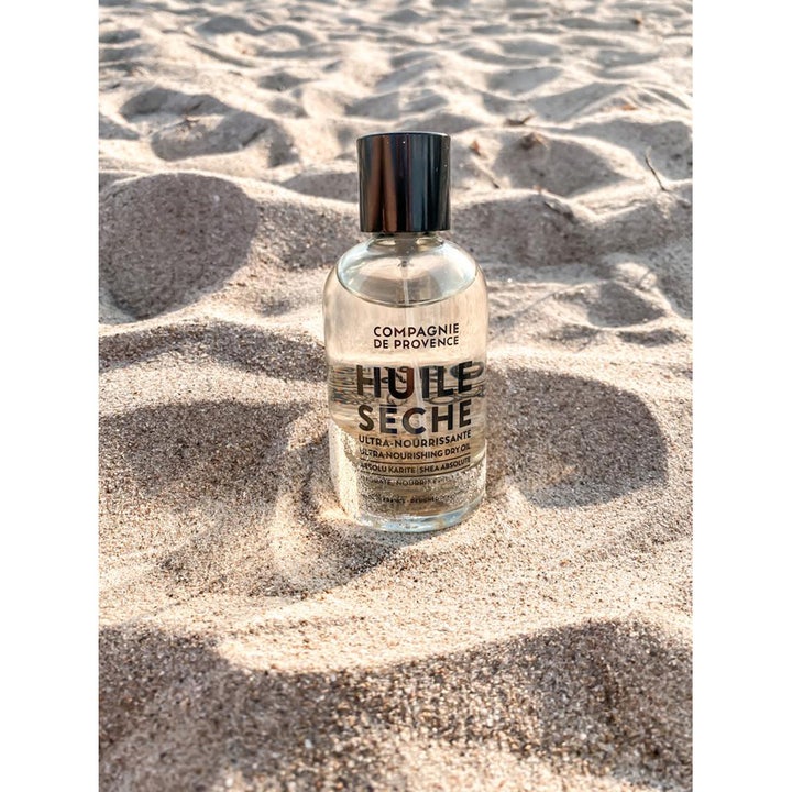 Compagnie de Provence Ultra Nourishing Dry Oil 100 ml kroppsolje i glassflaske på en solfylt sandstrand.