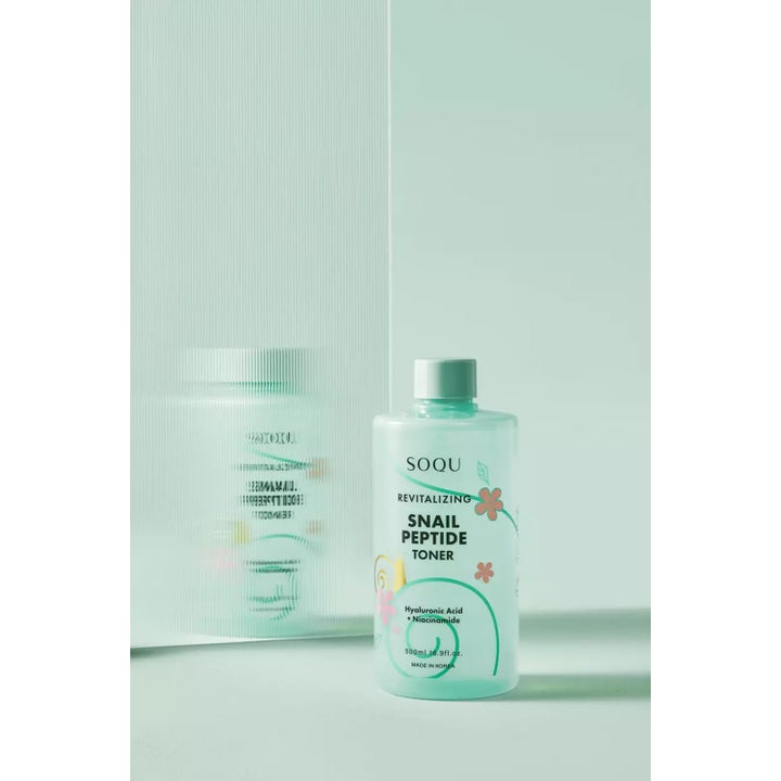 SOQU Snail Peptide Toner 500 ml revitaliserende toner i mintgrønn flaske med blomster ved en riflet glassvegg.