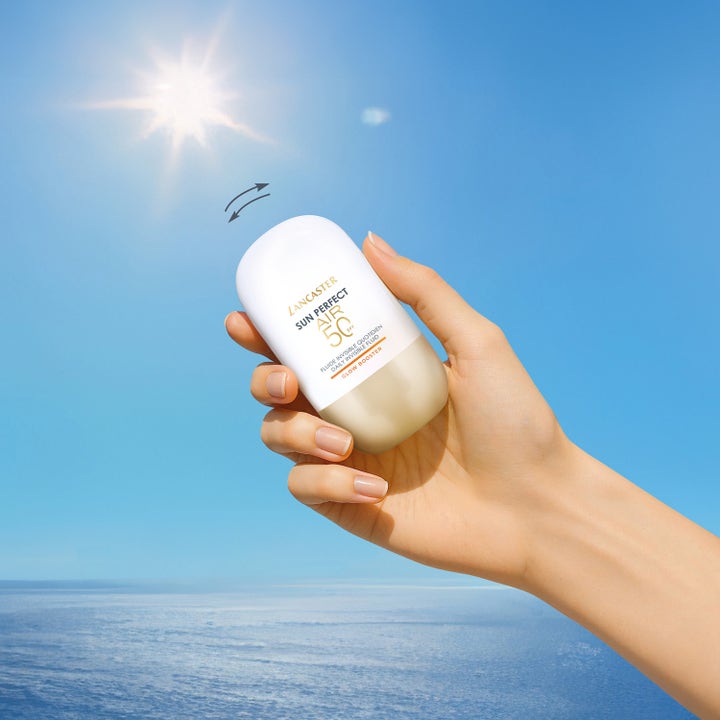 Lancaster Sun Perfect AIR Glow Booster SPF50 40 ml solkrem for ansiktet i en hånd mot solfylt himmel og hav.