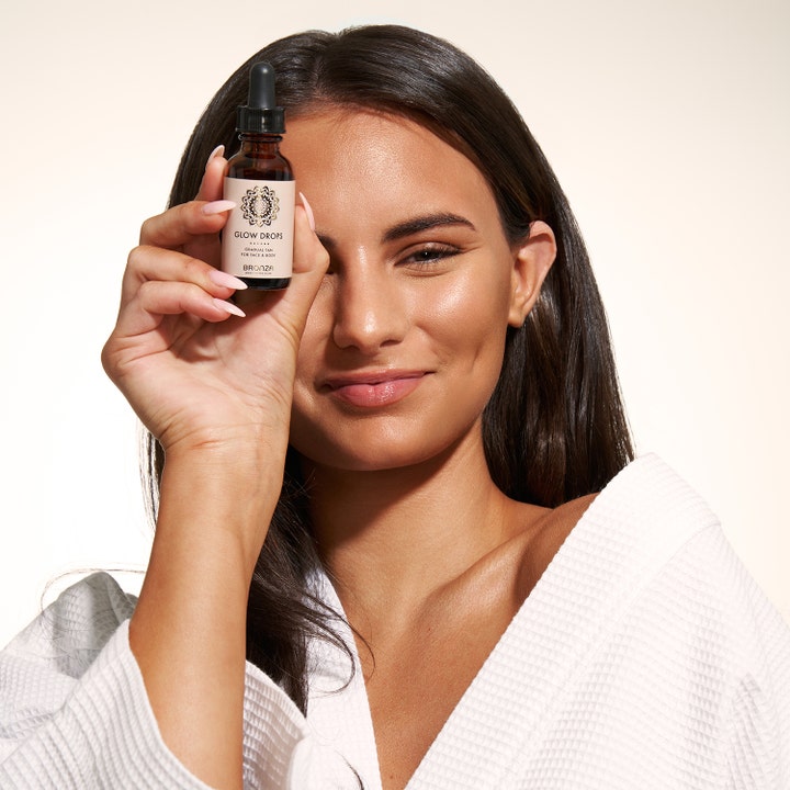 Bronza Glow Drops 30 ml tanning drops gir gradvis brunfarge vist av kvinne som holder mørk glassflaske med pipette