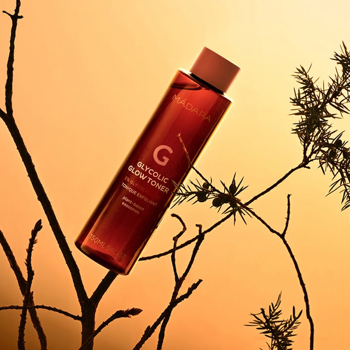 MÁDARA Glycolic Glow Toner 150 ml eksfolierende toner som gir glød i rød flaske plassert mellom greiner.