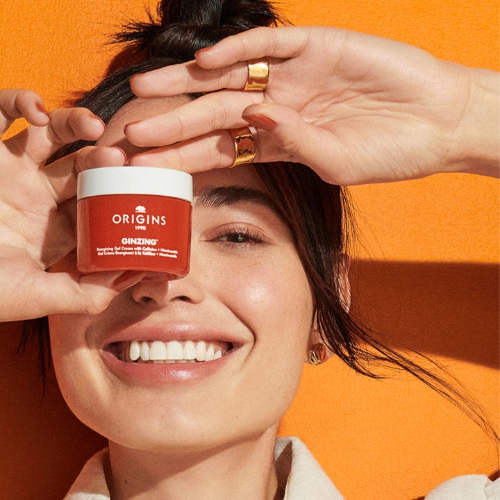 Origins Ginzing Energizing Gel Face Cream 50 ml gir glød i en oransje krukke som holdes av en smilende kvinne.