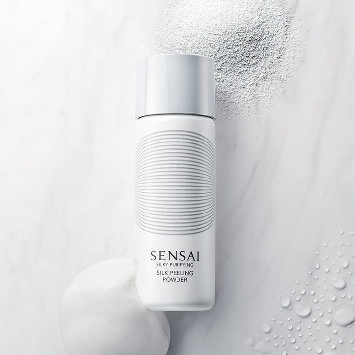 Sensai Silk Peeling Powder dyprensende ansiktsskrubb i hvit flaske med løst pudder og fyldig skum på lys marmor