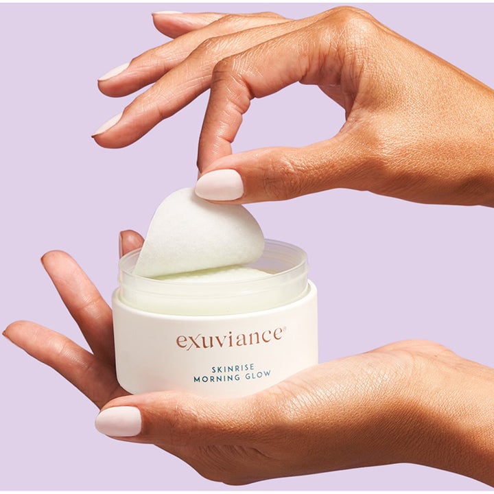 Exuviance Morning Glow 36 stk glødgivende pads i hvit krukke holdes av hender mot lilla bakgrunn